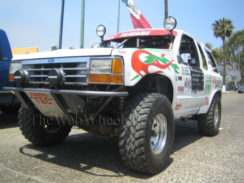1990 Ford Bronco - white lifted Ford Explorer race SUV with roof lights and colorful chili-pepper graphics sits on big tires near a Mexican flag.