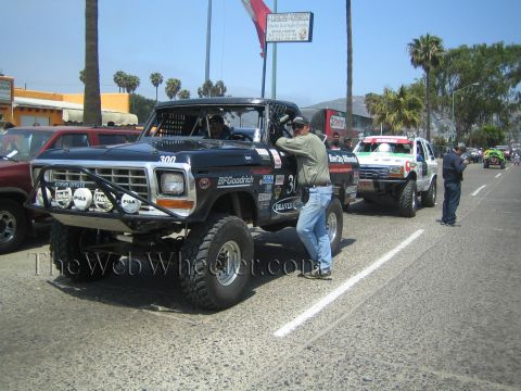 1990 Ford Bronco - black vintage Ford pickup race truck numbered 300 sits on a palm-lined street while a man in jeans leans into the window.