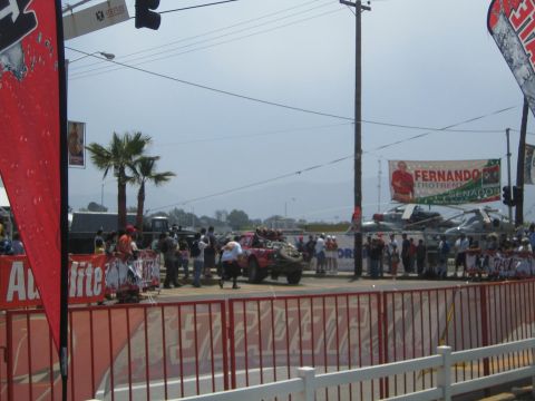 1990 Ford Bronco - off-road race truck stages at a start arch behind red banner barriers as a crowd gathers under a hazy sky.