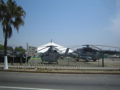 1990 Ford Bronco - Two gray military helicopters sit parked on a lot behind a chain-link fence, with a palm tree and tent in the background.