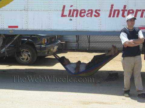 1990 Ford Bronco - man relaxes in a colorful hammock slung under a semi-truck trailer while a second man in a cap stands with arms crossed.