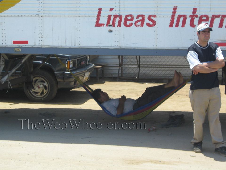 Man resting in a striped hammock under a semi trailer while another man stands beside it