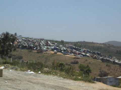 1990 Ford Bronco - sprawling hillside junkyard packed with rows of wrecked cars stretches across a dry landscape under a hazy sky.