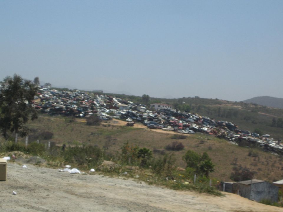 Distant hillside junkyard packed with rows of scrapped cars under a hazy blue sky