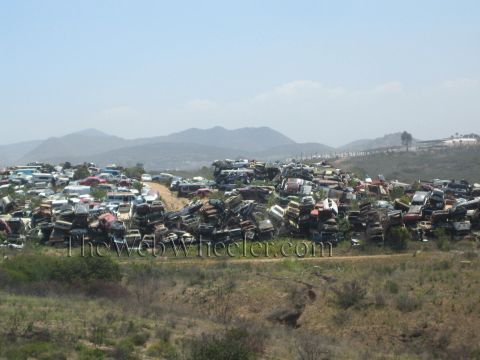 1990 Ford Bronco - Stacks of scrapped and rusting cars fill a hillside junkyard with distant mountains rising behind under a hazy sky.