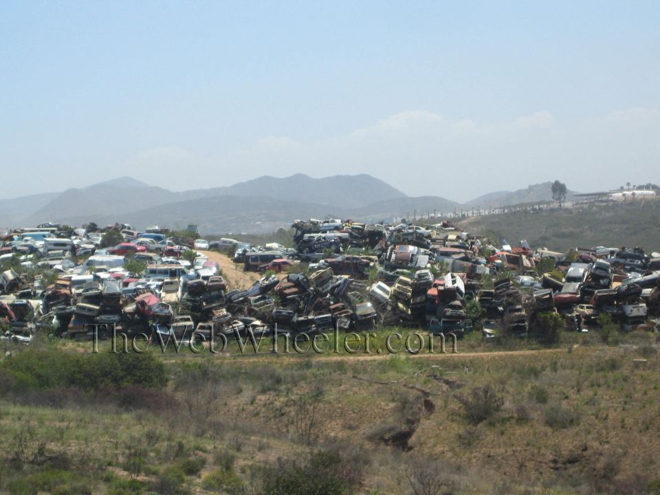 Large junkyard of stacked scrapped cars on a hillside with mountains in the background