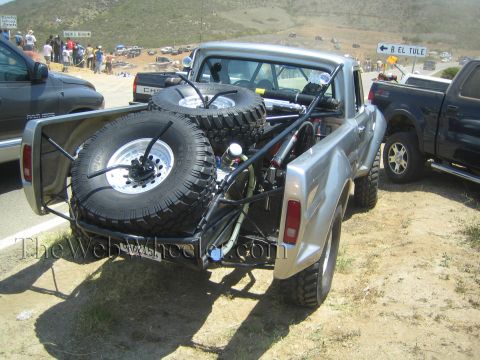 1990 Ford Bronco - Rear of a silver vintage Ford pickup prerunner with a tubular bed and two mounted spare tires, parked at an outdoor event.