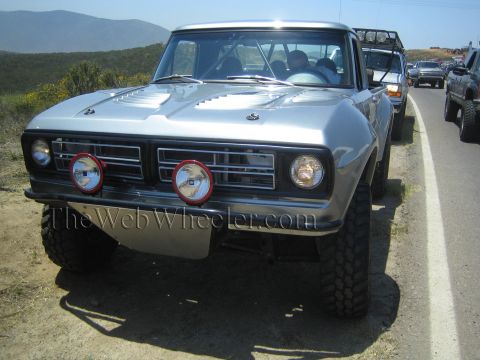 1990 Ford Bronco - Head-on view of a silver vintage Ford pickup prerunner fitted with two red-ringed round driving lights, parked on a hillside road.