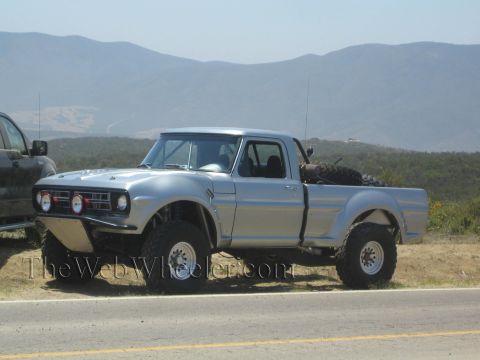 1990 Ford Bronco - silver vintage Ford pickup prerunner with round driving lights sits on a roadside shoulder with hazy mountains behind.