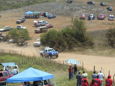 1990 Ford Bronco - blue off-road race truck throws up dust on a dirt course while spectators under tents watch from a hillside.