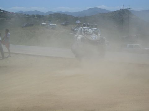 1990 Ford Bronco - silver off-road race buggy throws a thick cloud of dust on a dirt track as spectators and chase trucks watch from a hillside.