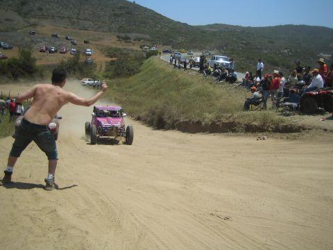 1990 Ford Bronco - pink single-seat off-road buggy races down a sandy hill while a shirtless spectator throws his arms up and crowds line the course.