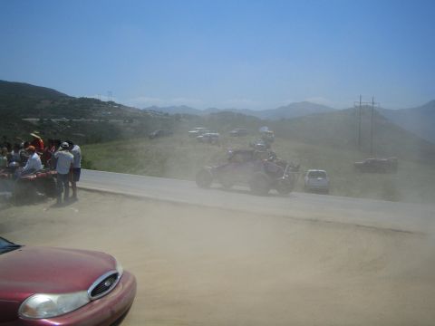 1990 Ford Bronco - off-road race buggy crosses a paved hilltop road in a haze of dust as spectators and a red car watch from the foreground.
