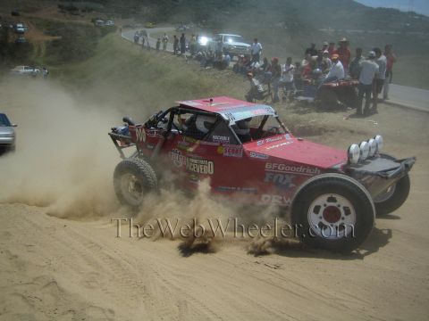 1990 Ford Bronco - red number 300 off-road race buggy plastered with BFGoodrich and Fox sponsor decals slides through a dusty dirt corner before a crowd.