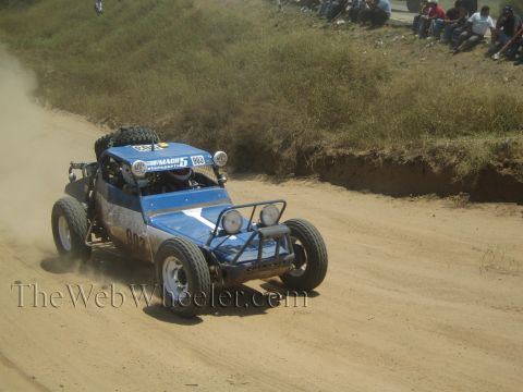 1990 Ford Bronco - blue and white tube-frame off-road race buggy speeds down a packed dirt road as spectators sit along the embankment above.