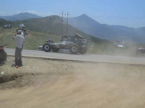 1990 Ford Bronco - camouflage-painted off-road buggy launches over a dirt berm beside a paved road as a helmeted spectator photographs it.