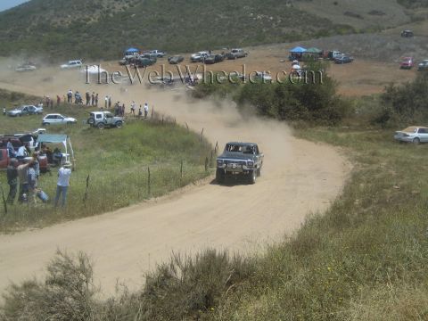 1990 Ford Bronco - light-colored off-road race truck rounds a sweeping dirt-road curve on a grassy hillside lined with spectators and parked support trucks.