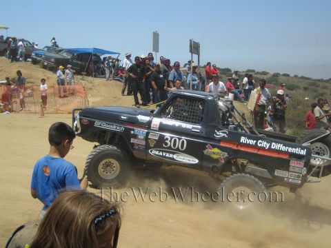 1990 Ford Bronco - black number 300 off-road race truck lettered River City Differential and BFGoodrich powers along a dirt course as a crowd looks on.