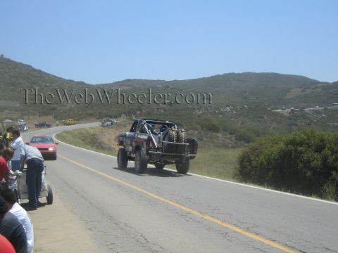 1990 Ford Bronco - lifted off-road race truck with a spare tire on its bed drives away down a paved hillside road as spectators watch from the shoulder.