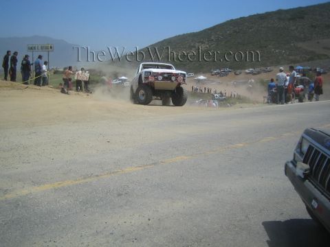 1990 Ford Bronco - light-colored off-road race truck catches air off a dirt rise next to a paved road, with spectators and a parked SUV framing the scene.