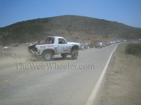 1990 Ford Bronco - white number 382 off-road race truck slides through dust on the dirt shoulder of a paved hillside road as cars line up behind.