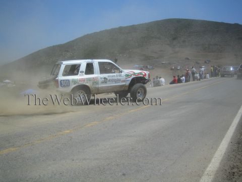 1990 Ford Bronco - white number 301 off-road race SUV with green and red striping drifts through dust along a paved hillside road past spectators.