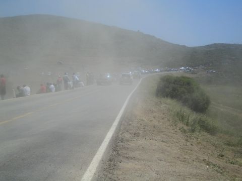 1990 Ford Bronco - paved hill road disappears into a haze of dust, lined with spectators and parked vehicles along a dry brushy slope.