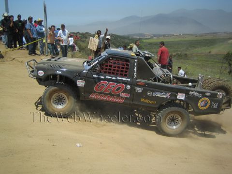 1990 Ford Bronco - black number 746 off-road race truck lettered GEO Ensenada slides up a dirt rise as a large crowd cheers from the hillside.