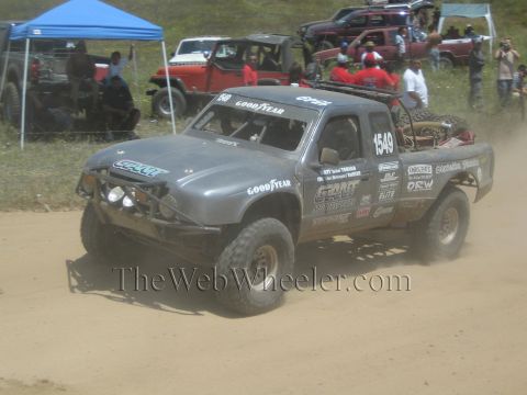 1990 Ford Bronco - gray number 1549 off-road race truck with Goodyear and Grant decals drives a dirt course past blue pop-up tents and onlookers.