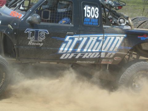 1990 Ford Bronco - Close-up of a black number 1503 race truck door reading Stinson Offroad and LTS Racing as the rear tire throws up dust.