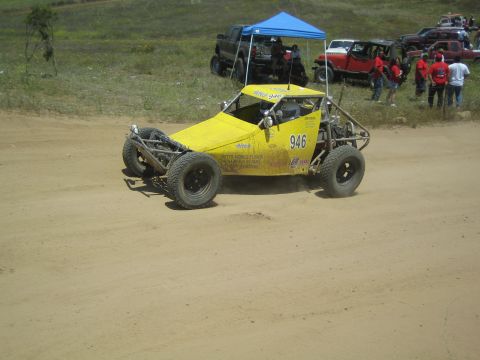 1990 Ford Bronco - yellow number 946 tube-frame off-road buggy sits on a sandy course near a blue pop-up tent and a row of standing spectators.