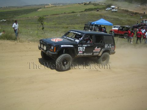 1990 Ford Bronco - black number 1702 Jeep Cherokee JeepSpeed race SUV corners on a dirt course before a green hillside and spectators with tents.
