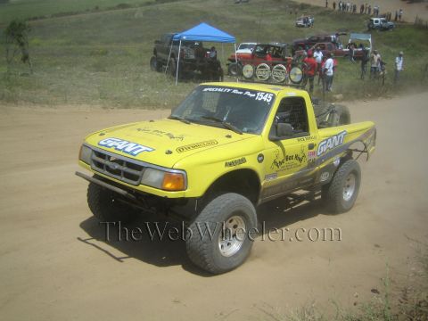 1990 Ford Bronco - yellow number 1545 off-road race truck lettered GIANT sits on a sandy course with pop-up tents and parked rigs in the background.