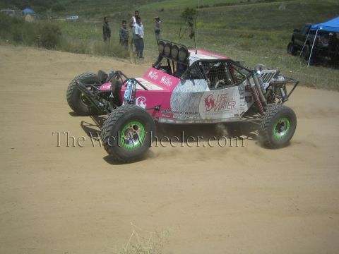 1990 Ford Bronco - pink and white tube-frame race buggy with bright green wheels and Life Water decals slides through a dirt corner past spectators.