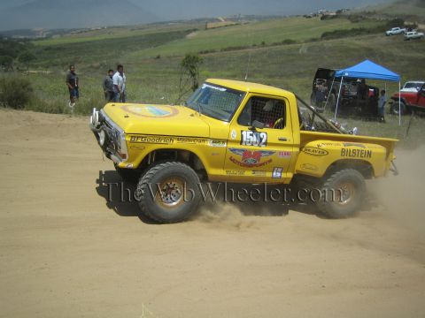 1990 Ford Bronco - yellow Ford number 1542 off-road race truck with BFGoodrich and Bilstein decals slides across a dirt course on a green hillside.