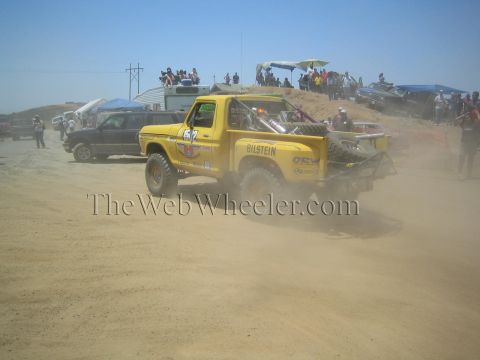 1990 Ford Bronco - yellow Ford off-road race truck with Bilstein decals throws a heavy dust cloud on a sandy course packed with spectators.