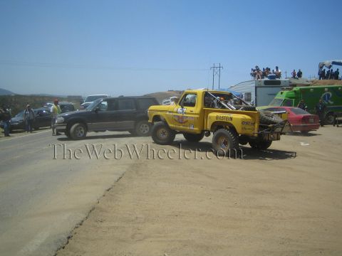 1990 Ford Bronco - yellow Ford off-road race truck with Bilstein decals waits on a dirt road behind a dark pickup as spectators gather nearby.