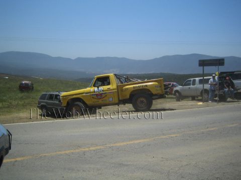 1990 Ford Bronco - yellow Ford number 1542 off-road race truck sits on a paved roadside with crews and chase vehicles against a mountain backdrop.