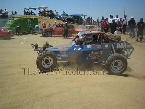 1990 Ford Bronco - blue single-seat off-road race buggy numbered 948 kicks up dust on a dirt course as a crowd of spectators and an ambulance watch from the h…