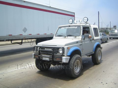 1990 Ford Bronco - silver lifted Suzuki SUV with a brush guard, roof-mounted lights and blue fender flares drives a highway past a semi trailer.
