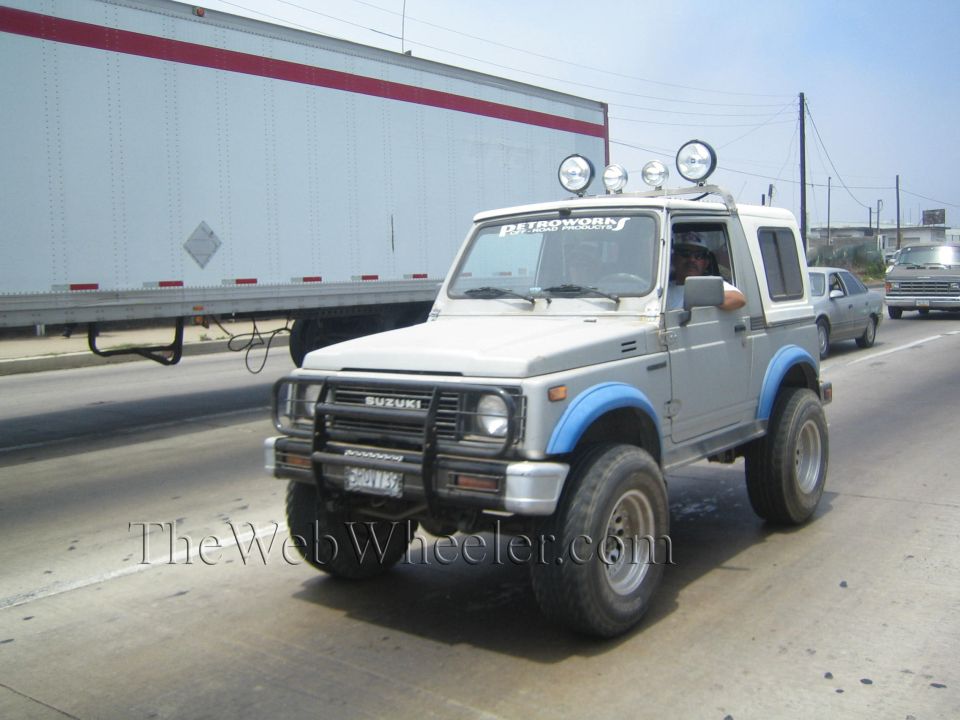 Silver Suzuki SUV with roof lights and blue fenders driving on a highway