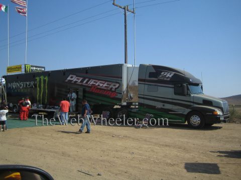 1990 Ford Bronco - black Monster Energy and Pfluffer Racing semi truck hauler sits at a dusty desert race pit as crew and spectators gather nearby.