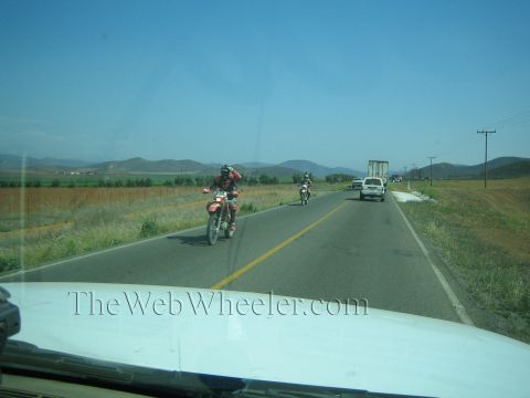 1990 Ford Bronco - Two dirt bike riders travel along a rural two-lane highway flanked by fields and hills, seen through a vehicle windshield.