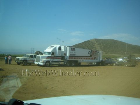 1990 Ford Bronco - white semi truck pulling a race-team trailer parks on bare dirt beside a desert hill as people and trucks gather around.