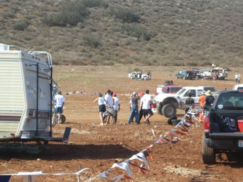 1990 Ford Bronco - Spectators walk among pickups and a parked travel trailer at a dusty off-road race camp, with bunting strung in the foreground.