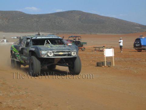 1990 Ford Bronco - dark Monster Energy Chevrolet trophy truck races across red desert dirt past a course marker and parked vehicles.