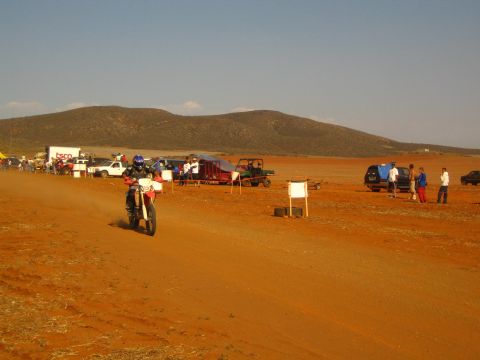 1990 Ford Bronco - helmeted rider speeds along a red-dirt desert course on a dirt bike past spectators, trucks and course markers.