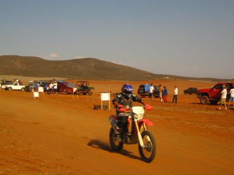1990 Ford Bronco - helmeted rider in blue and black gear races a red Honda dirt bike across a red-dirt desert course with onlookers behind.