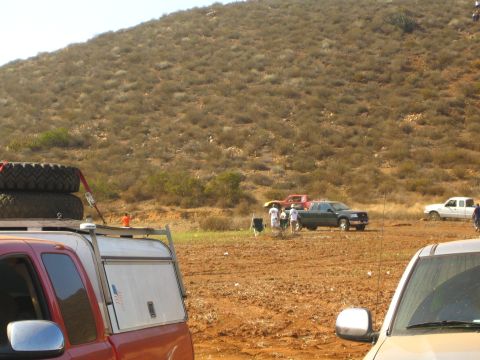 1990 Ford Bronco - Several pickups, one carrying a spare tire, are parked on bare dirt as people gather near a brushy hillside off-road area.