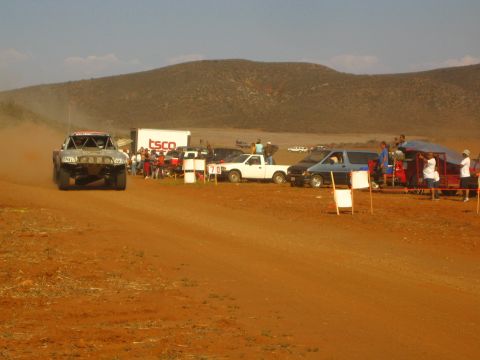 1990 Ford Bronco - off-road race truck throws up dust along a red-dirt course as spectators and parked pickups line the roadside below a hill.